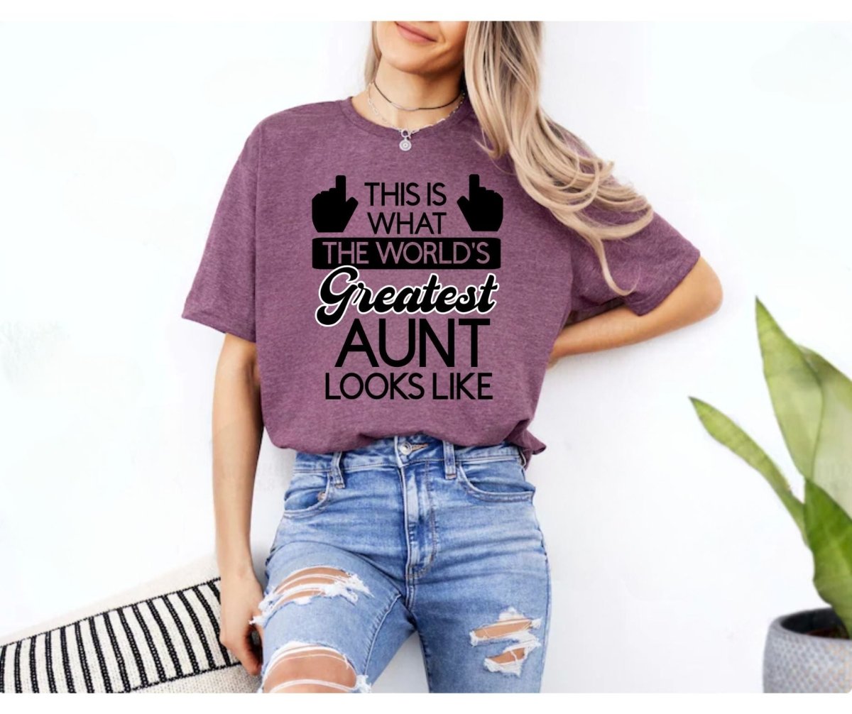 Woman standing with World's Greatest Aunt plum color heather maroon unisex tshirt with black and white lettering on. This is what the world's greatest aunt looks like on front with hands pointing up to the person wearing the shirt.. Little Gift Nook.