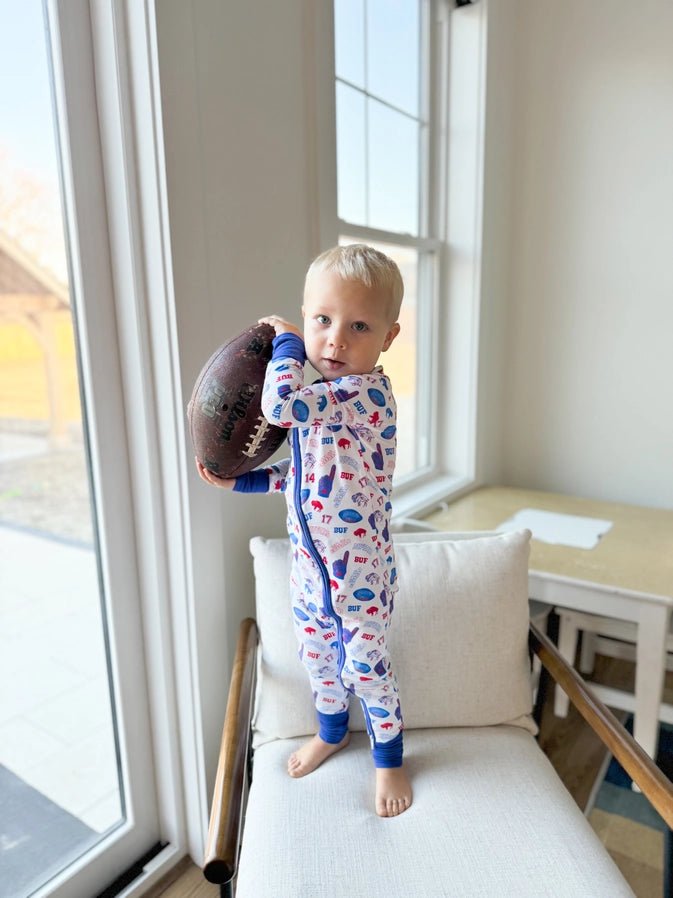Toddler standing holding a football in bamboo buffalo football print #17 and #86 royal blue sleeper standing on chair.