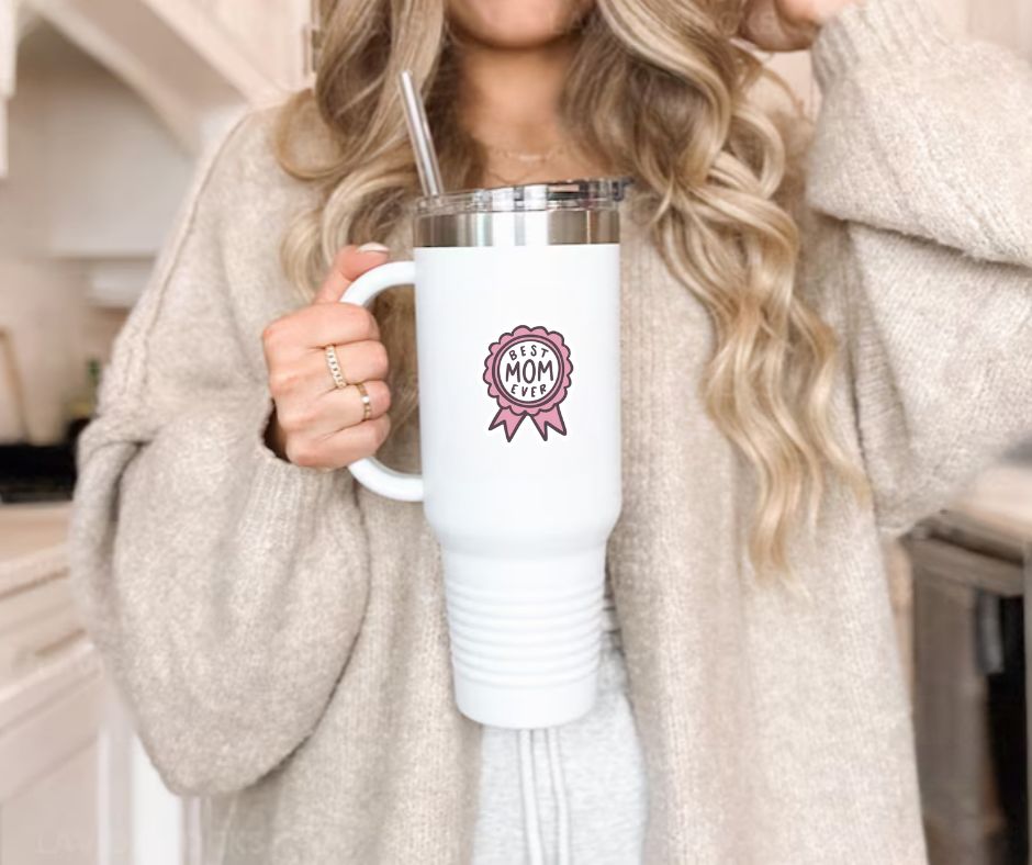 Woman holding white tumbler with pink ribbon image sticker with "best MOM ever" on front sitting on gray background. Little Gift Nook