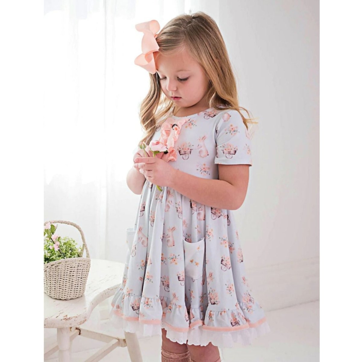 Honey Bunny Blue Eyelet Twirl Dress. Light baby blue dress with pink bunnies and ruffle accents. Eyelet trim and 2 front pockets. Little girl wearing dress holding flowers. - Little Gift Nook