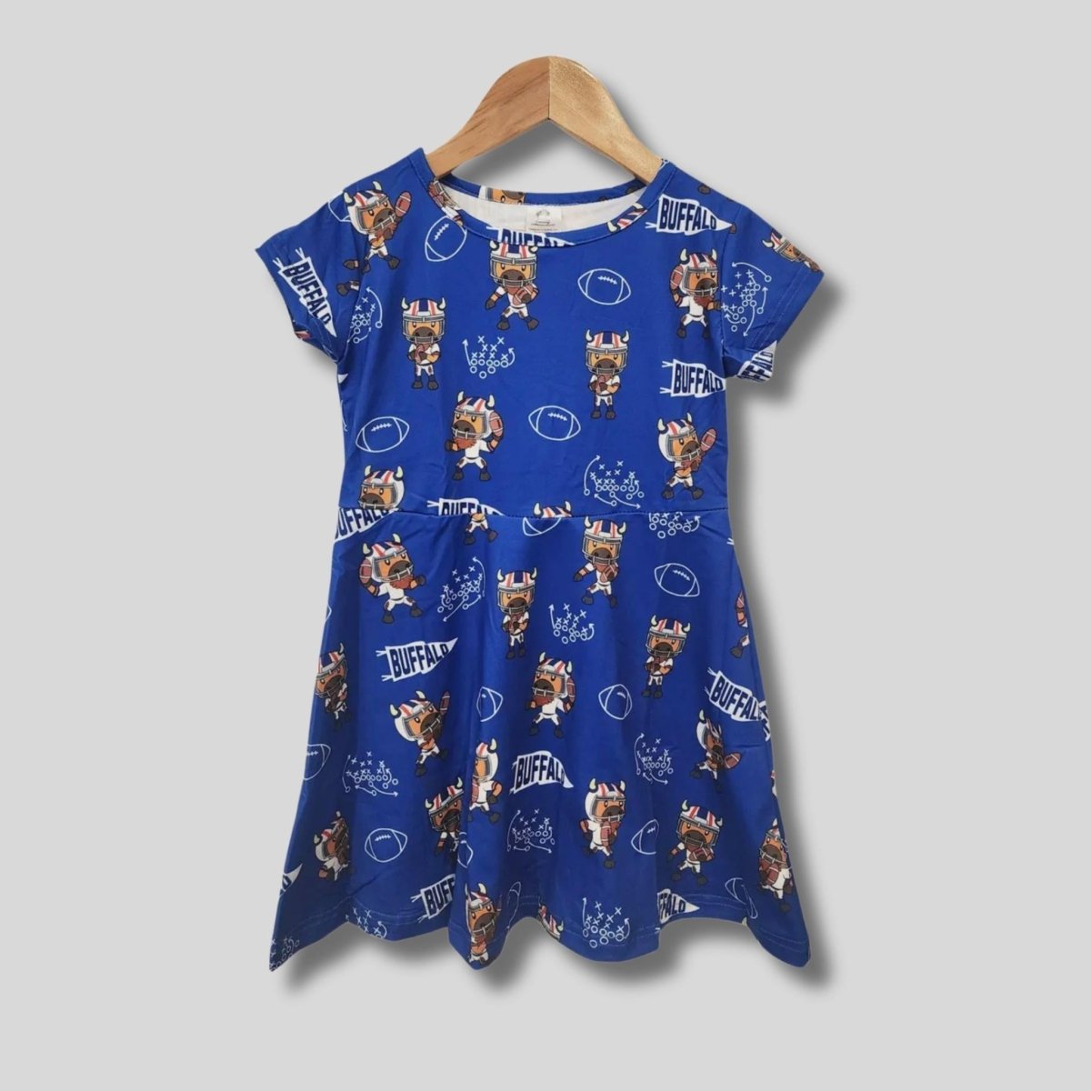 Royal blue girl's quarterback cartoon buffalo football print dress hanging on wooden hanger on gray background.