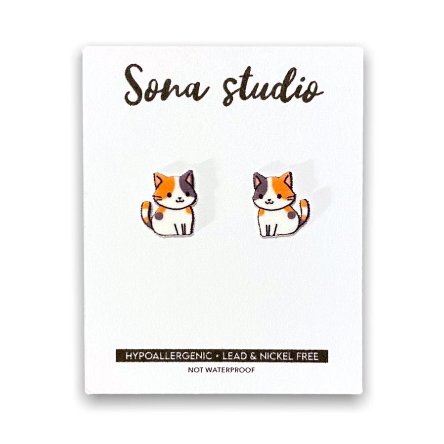 White, orange, and gray calico cat earrings. Cartoon image handmade stud earrings on a white cardstock. Little Gift Nook.