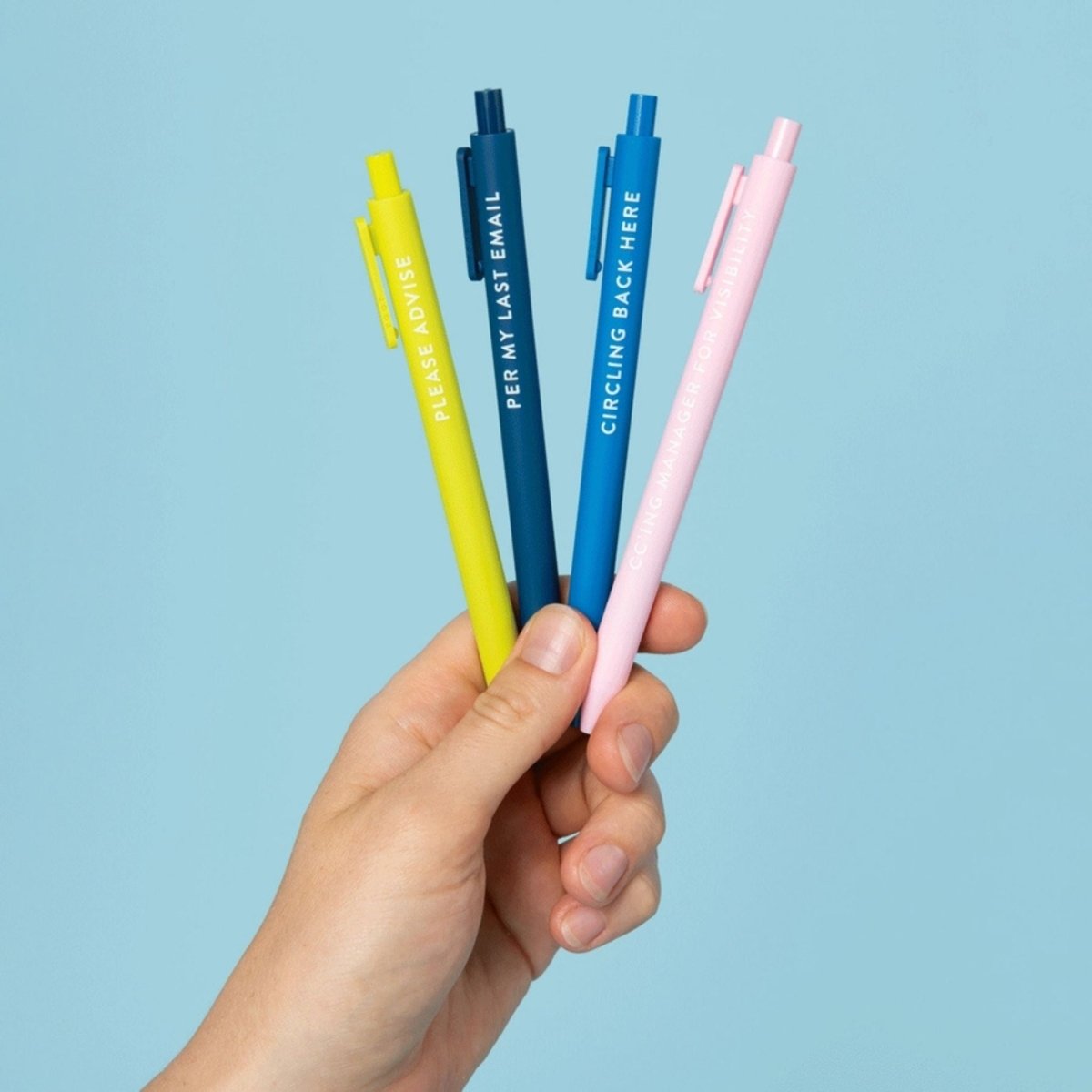 4 pens in green, navy, blue, and pink with funny corporation passive aggressive sayings on them being held up by a hand on blue background.