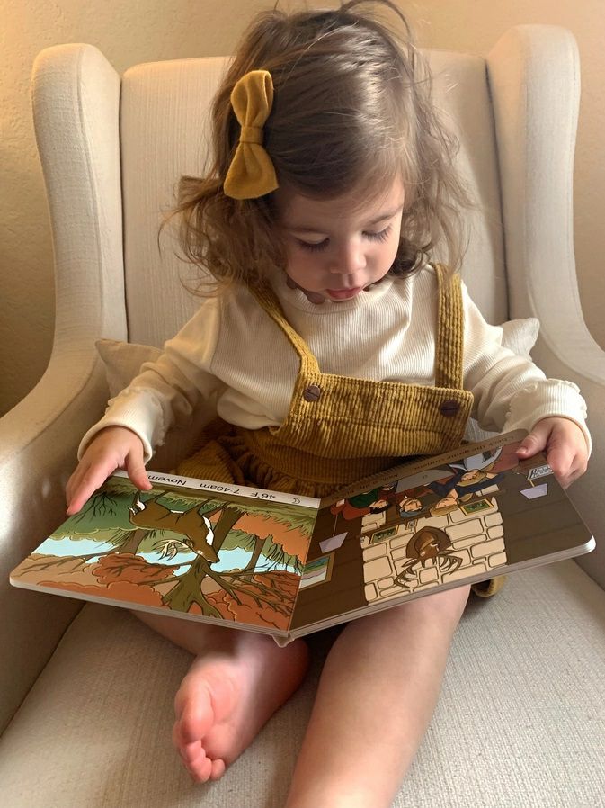 Little girl reading Deer Camp board book sitting in tan chair.