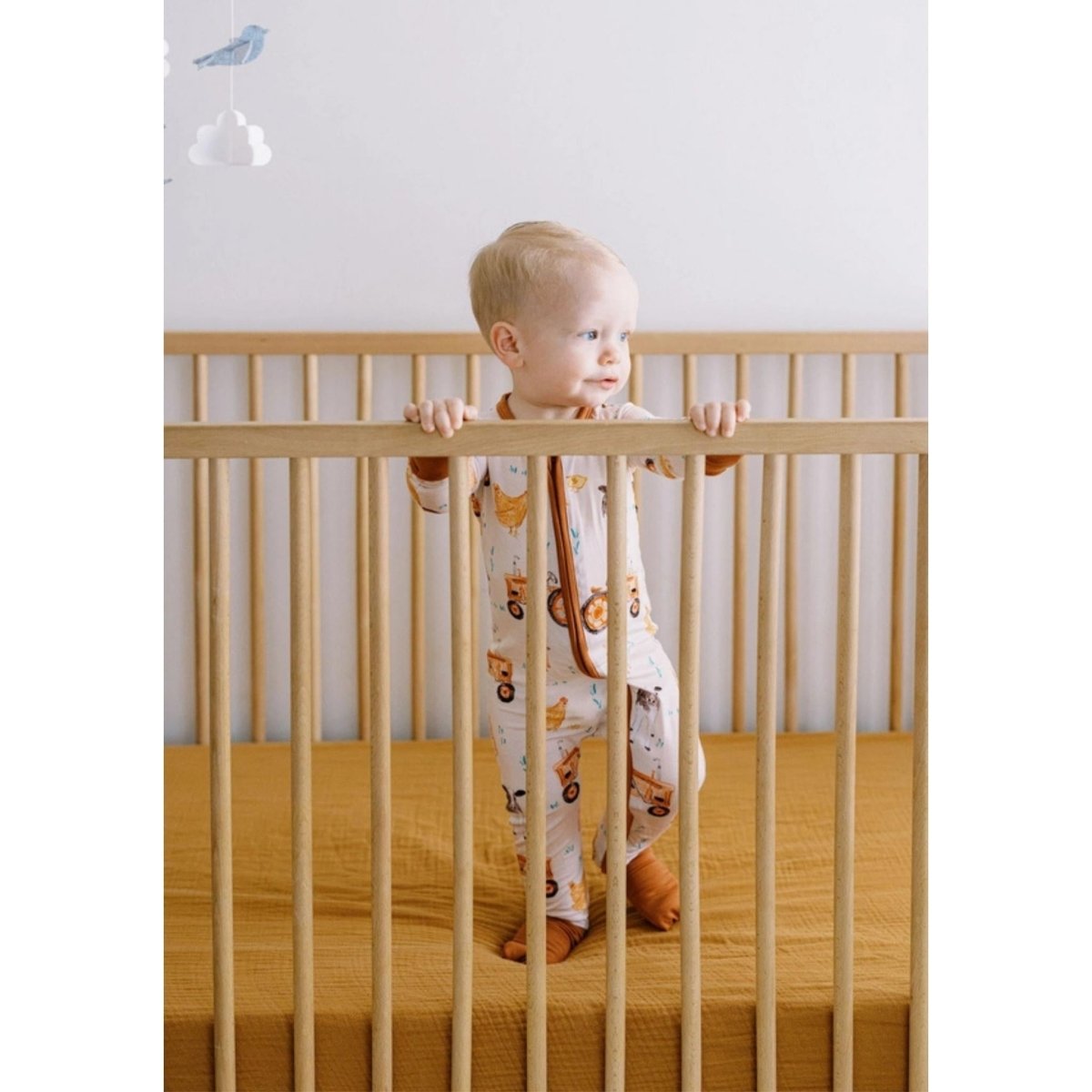 Baby standing in crib wearing brown farm theme bamboo sleeper.