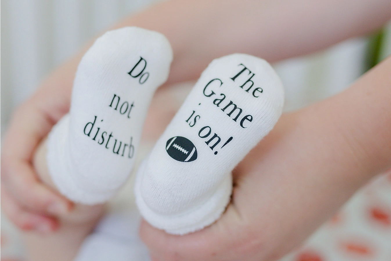 white baby socks with "Do not disturb" "The Game is on" written on bottoms on baby's feet behind held up.