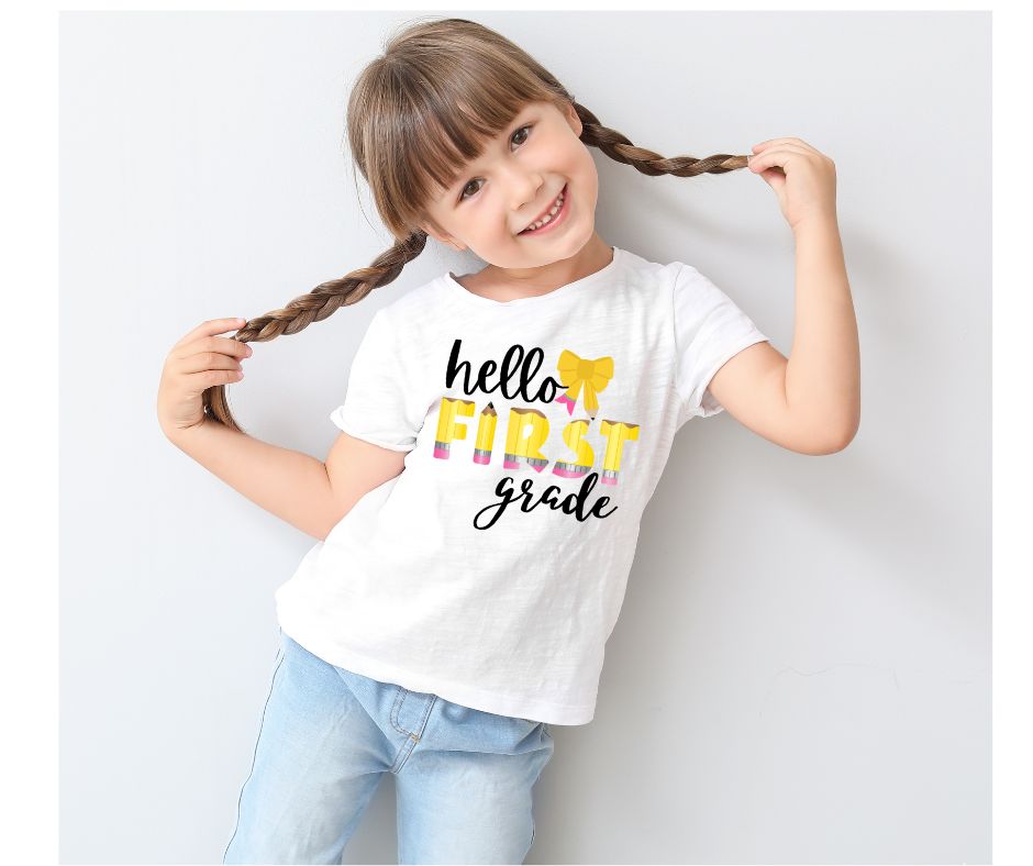 Child wearing a 'hello FIRST grade' t-shirt with the word FIRST in a pencil theme and a yellow bow. Digital image on white tshirt.