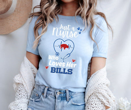 woman wearing a light blue t-shirt with a graphic and text "just a nurse who loves her bills" a stethoscope with red buffalo and heartbeat, standing against a neutral background.
