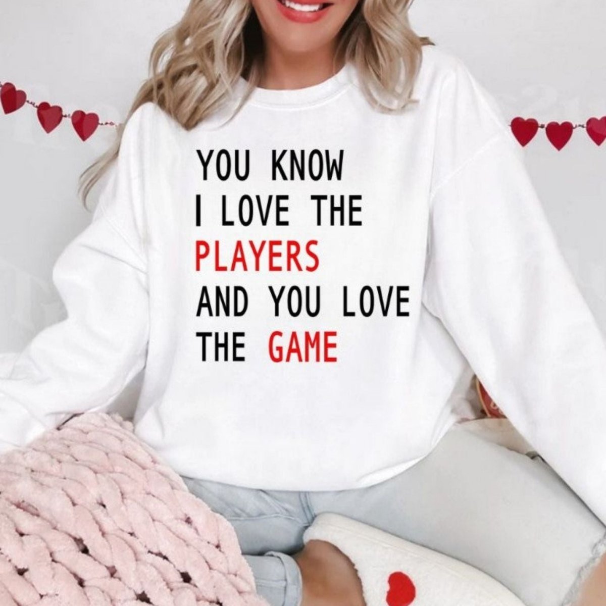 KC Love the Players Love the Game Sweatshirt. White fleece lined crewneck unisex sweatshirt with black and red play on lyrics sweatshirt on model with hearts in background on model sitting with shirt on and hearts in background. Little Gift Nook.