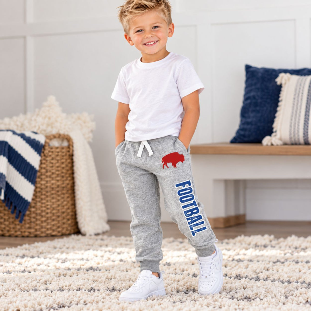 A young boy stands in a bright, modern room wearing heather gray sweatpants with a red buffalo graphic and the word 'FOOTBALL' in blue, paired with a white t-shirt and white sneakers.