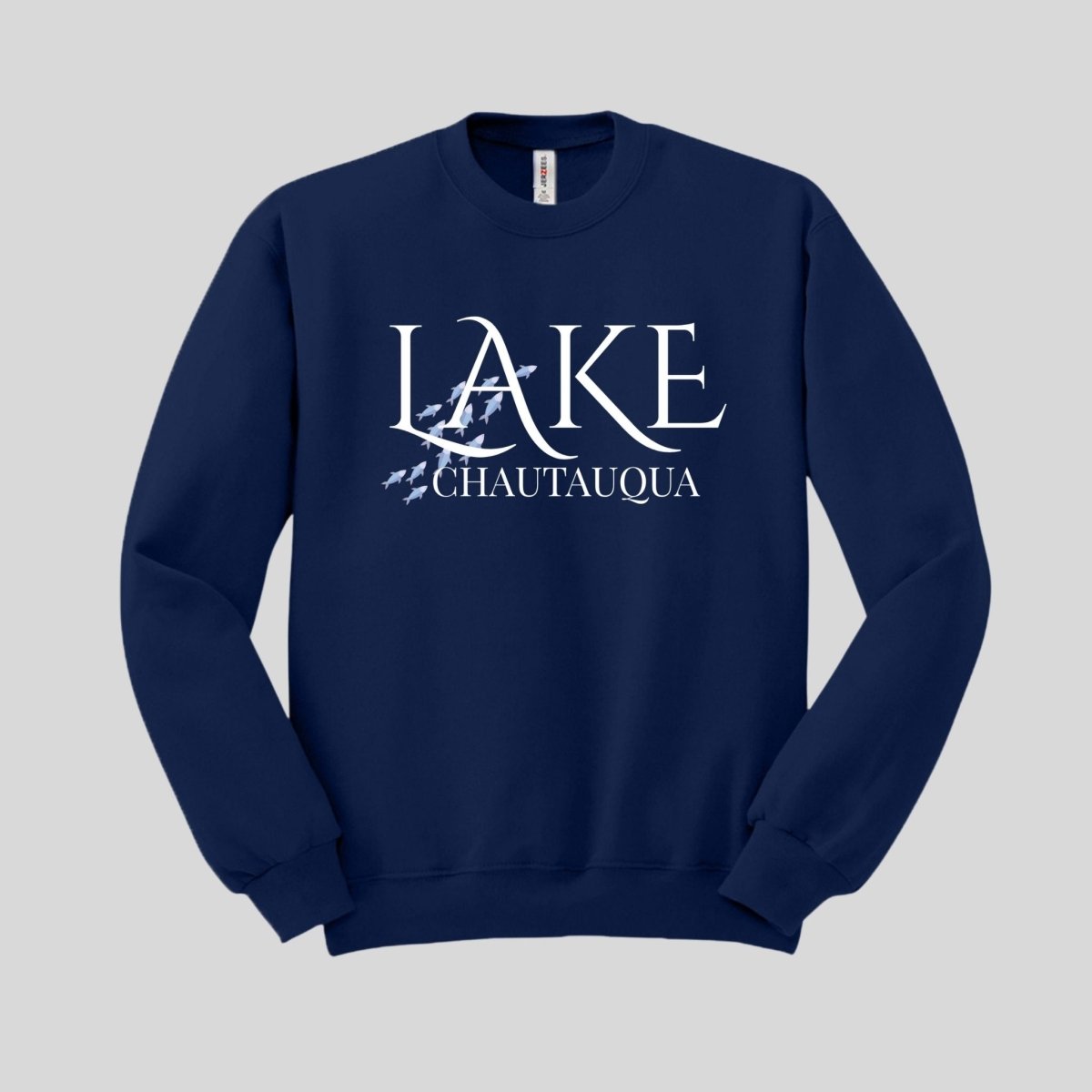 Navy sweatshirt with white text "LAKE CHAUTAUQUA" and blue fish swimming graphic on front laying on gray background. Little Gift Nook
