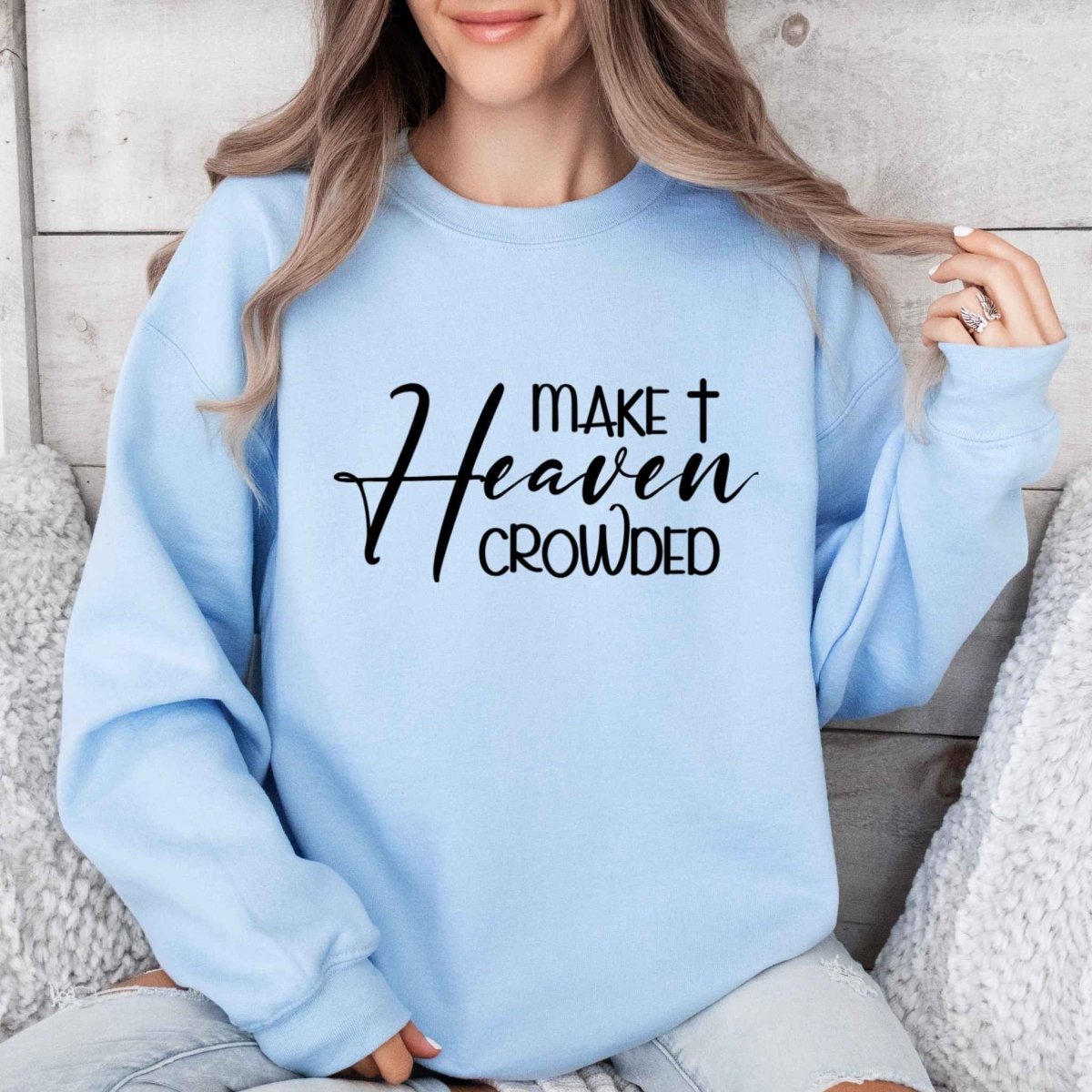 woman in baby blue crewneck sweatshirt with "Make Heaven Crowded" in black text on front on a sofa and gray background.