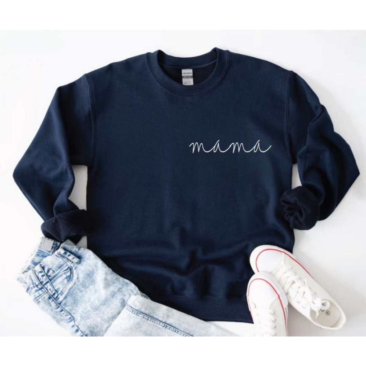 Navy crewneck sweatshirt with white mama on top left on a white background next to a pair of jeans and sneakers.