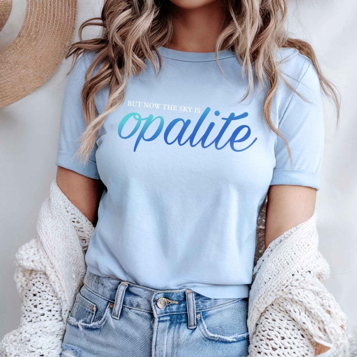 woman standing in light blue tshirt with white "BUT NOW THE SKY IS" AND "OPALITE" in Iridescent blue lettering on front on light gray background.