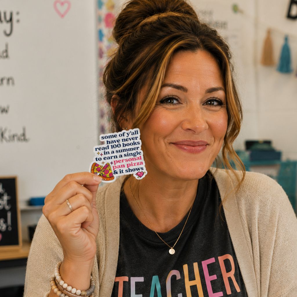 A smiling woman in a classroom setting holds up a small sticker with a humorous quote "some of ya'll have never read 100 books in summer to earn a single personal pan pizza and it shows" She is wearing a beige cardigan over a black graphic t-shirt that says 'TEACHER' in colorful letters.