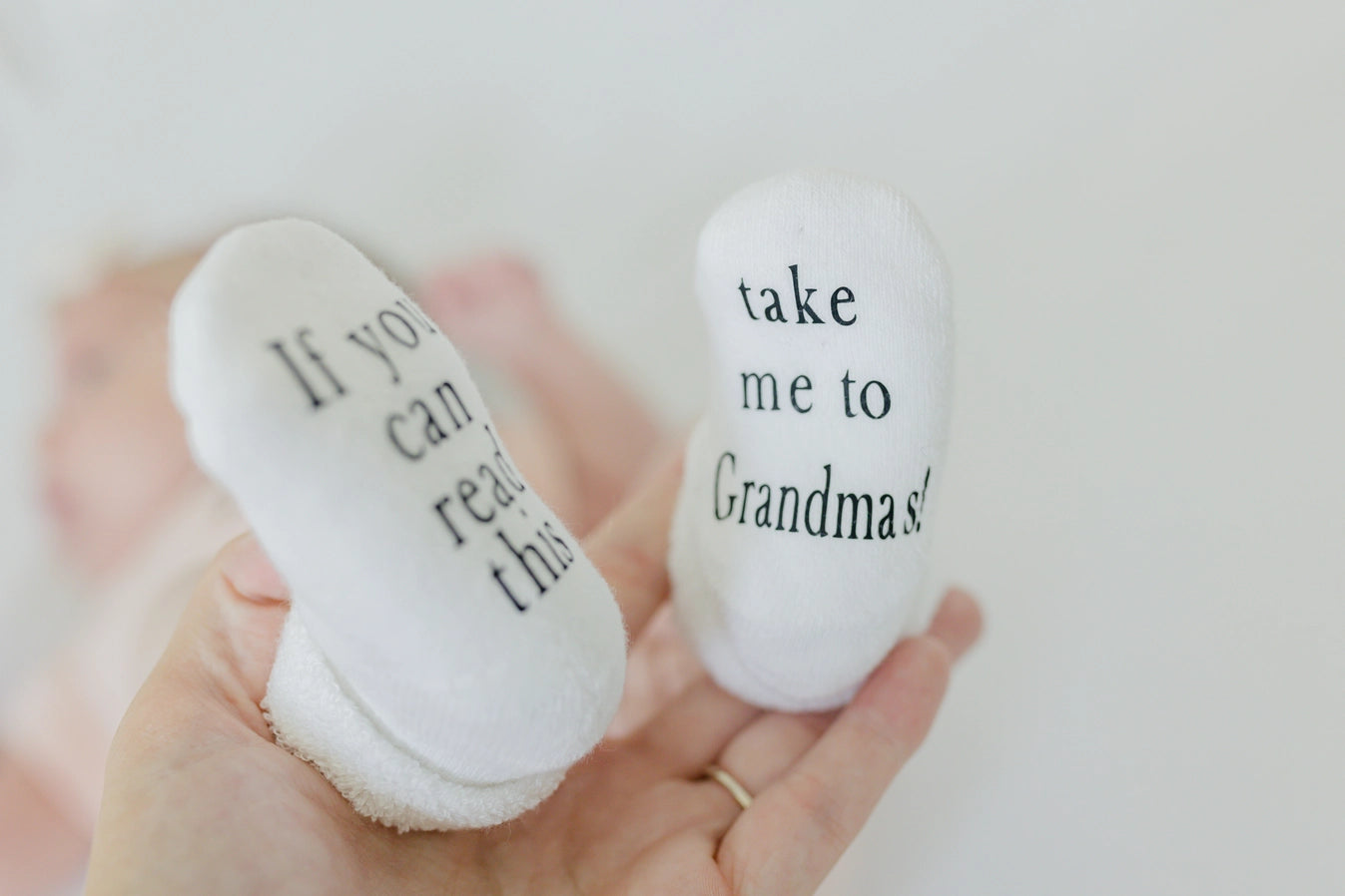 white baby socks on baby feet with text on bottoms "If you can read this" "take me to Grandma's" being held up by woman's hand.
