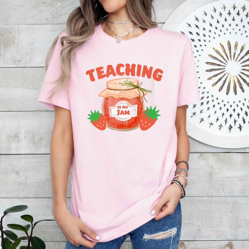 Woman wearing pink tshirt with orange/red jam jar, strawberries, and "Teaching is my jam" on front on a wood plank background. Little Gift Nook