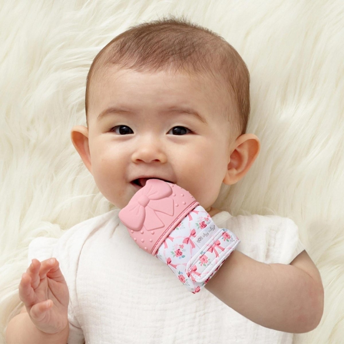 Baby chewing on Itzy Mitt wearable pink bow silicone teething mitt teether laying on faux fur background.
