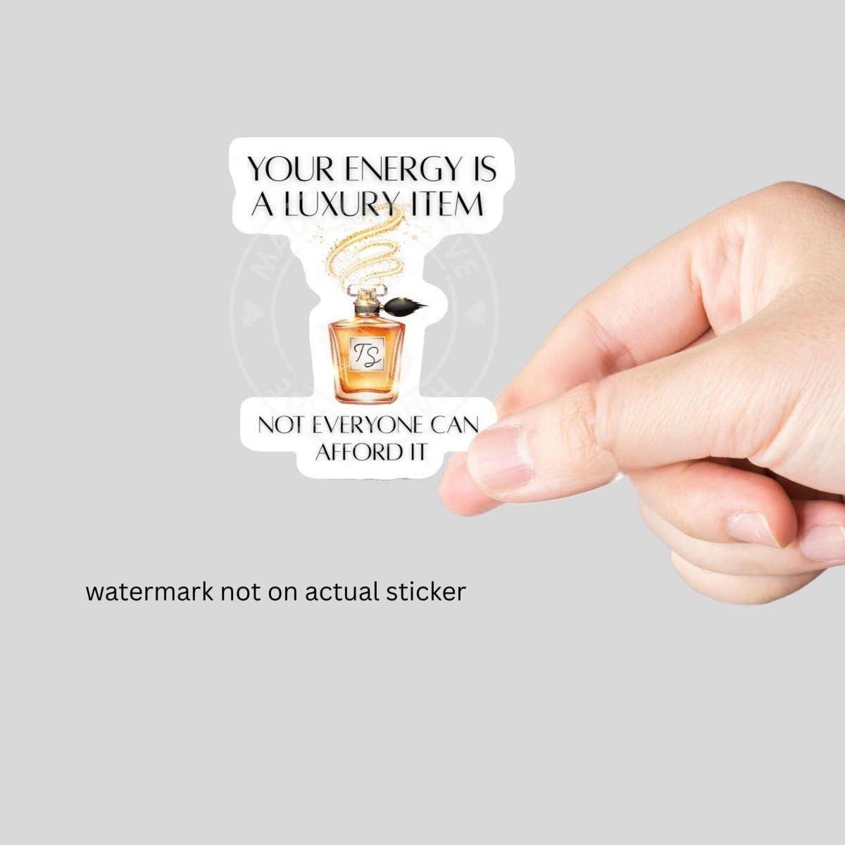 Orange perfume bottle "Your energy is a luxury item not everyone can afford it" sticker with white background held in a hand on a gray background.