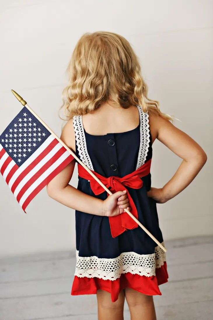 Back view of girl wearing navy, red, and white patriotic flag dress for kids, featuring button detail and red sash bow, holding American flag—Fourth of July children’s boutique outfit from Little Gift Nook