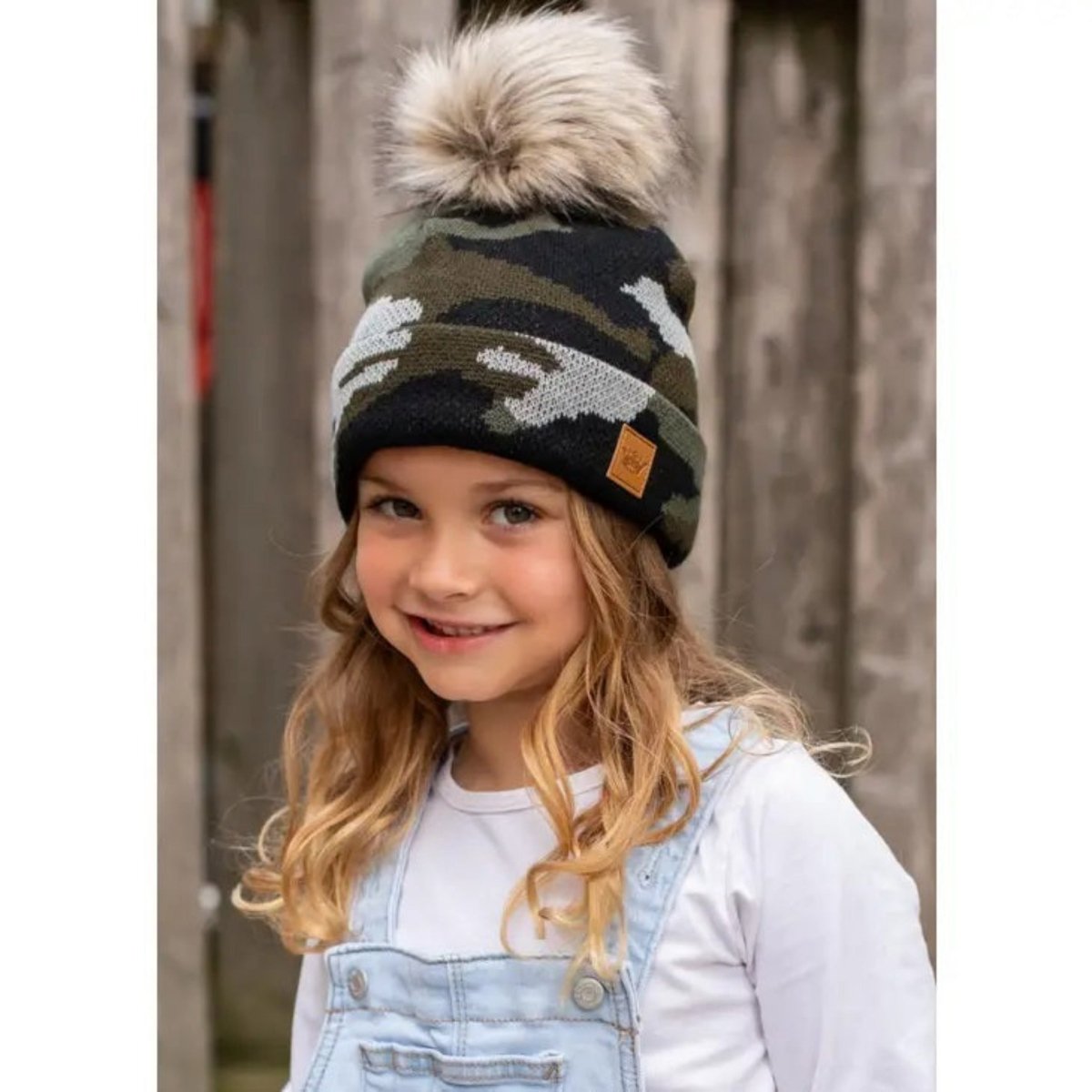 Little girl wearing green camo camouflage knit winter pom hat standing in front of wooden fence. Little Gift Nook