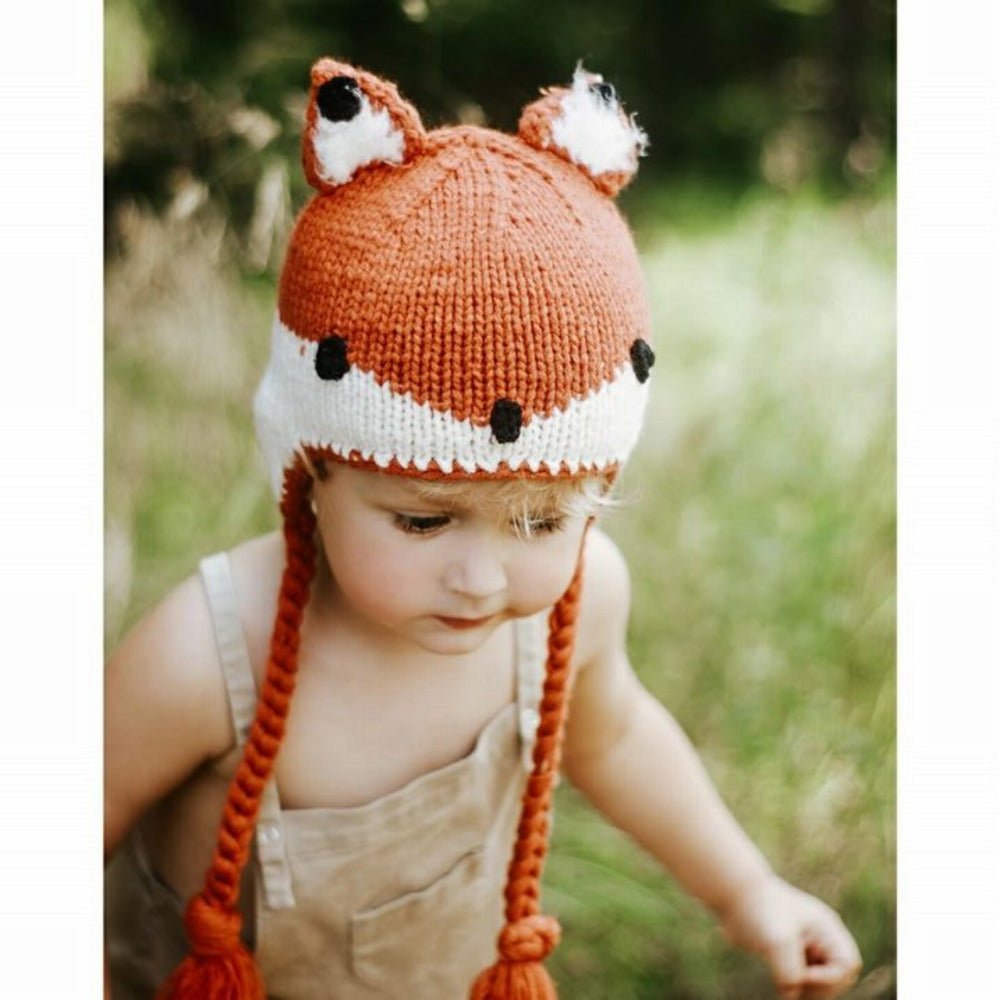 Toddler wearing orange fox knit hat with braided tassels walking in field.