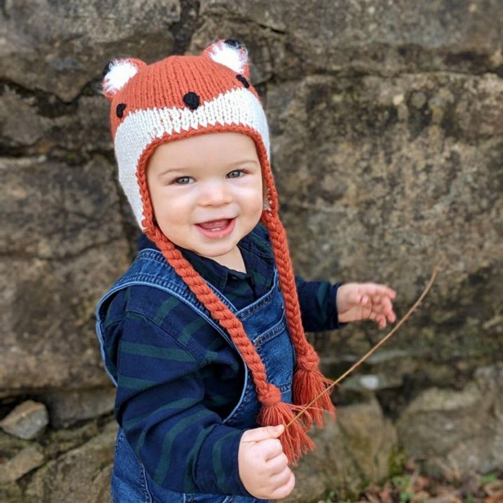 Toddler Boy wearing orange knit fox hat with braided tassels smiling standing next to rocks.