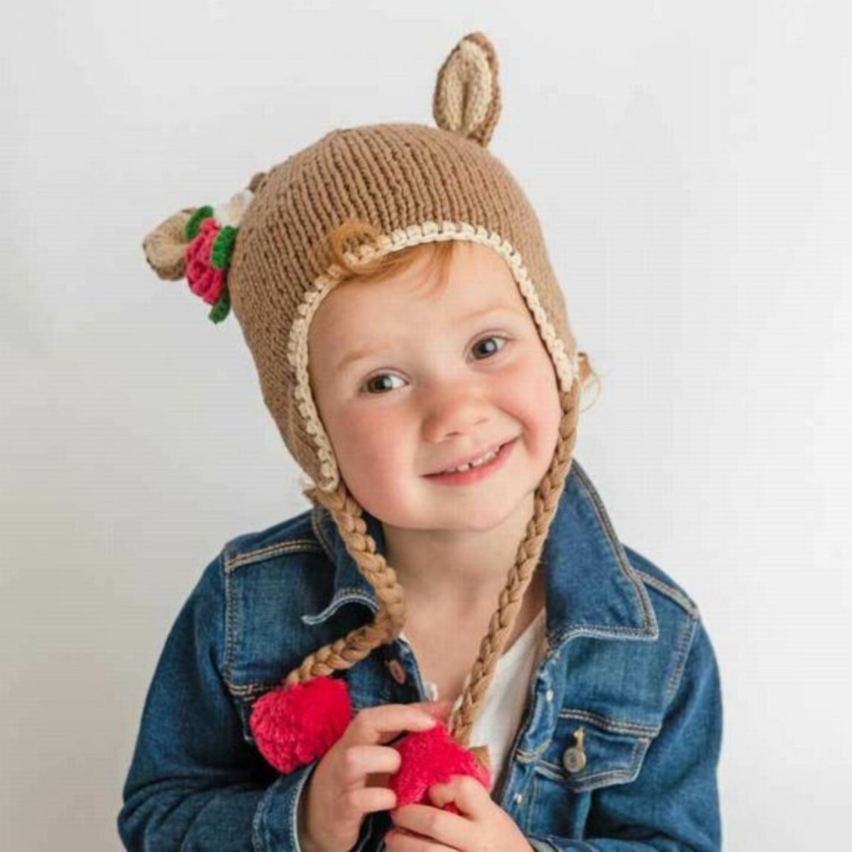 Little girl in knit deer doe hat with braided tassels and pink pom poms standing in front of a beige background.