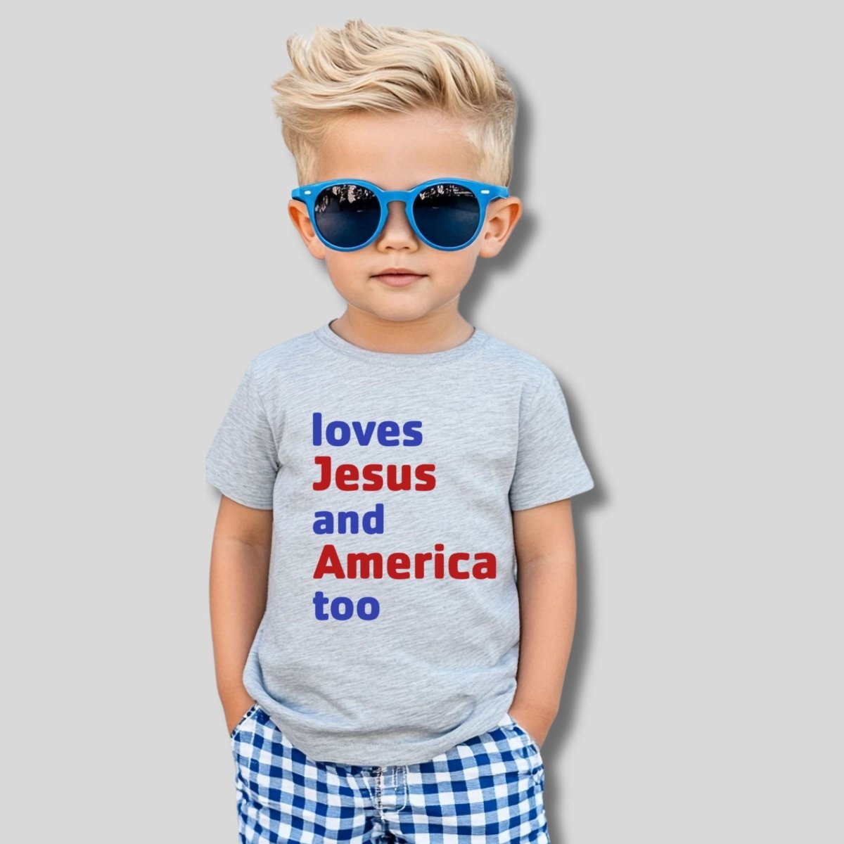 Little boy wearing gray tshirt that says "loves Jesus and America too" in red and blue text, wearing sunglasses, on a gray background.