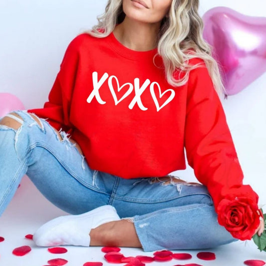 Red XOXO Puffy Letters Crewneck. Sweatshirt on model paired with ripped denim jeans. Unisex fit fleece lined red crewneck sweatshirt with white textured puffy lettering xoxo on front. Makes cute gift. Little Gift Nook.