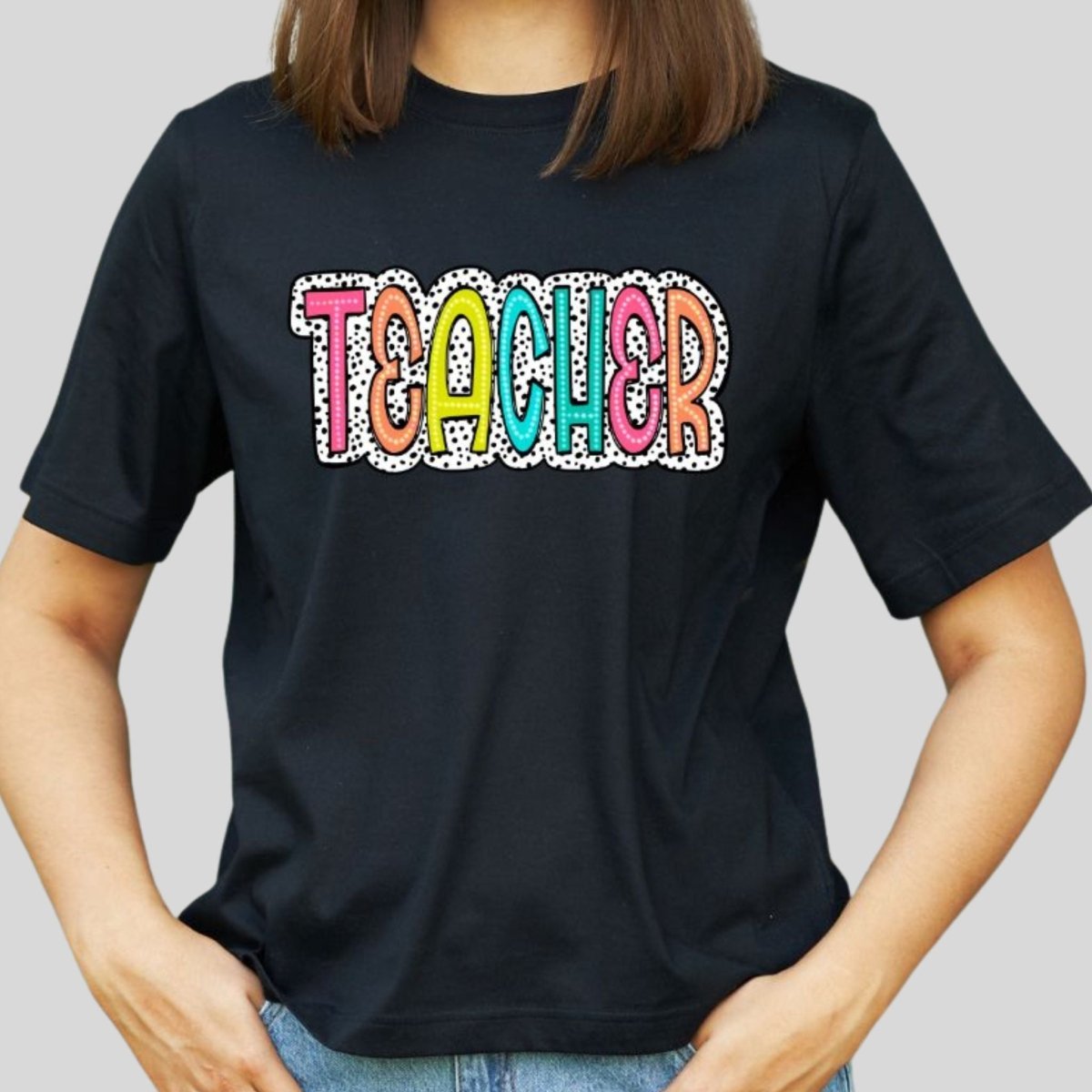 Woman wearing black short sleeve tshirt with colorful TEACHER on front and black white polka dot dalmatian print on front. Little Gift Nook.