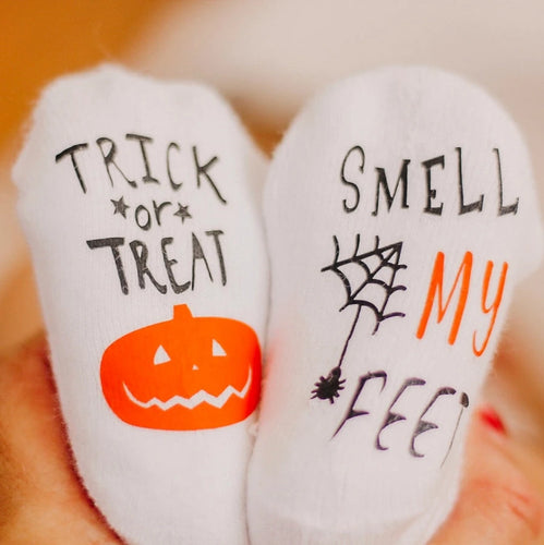 Trick or Treat Smell My Feet Baby Socks. White socks with black & orange lettering, spiderweb, and pumpkin on bottoms.