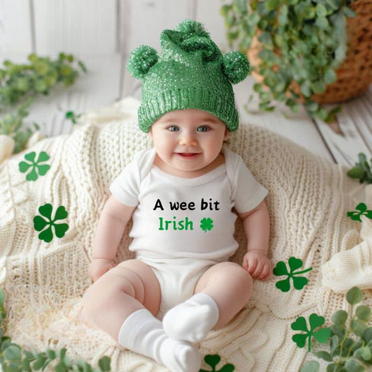 Baby wearing green hat and white bodysuit with "a wee bit Irish" on front and clover on a clover blanket background. Little Gift Nook