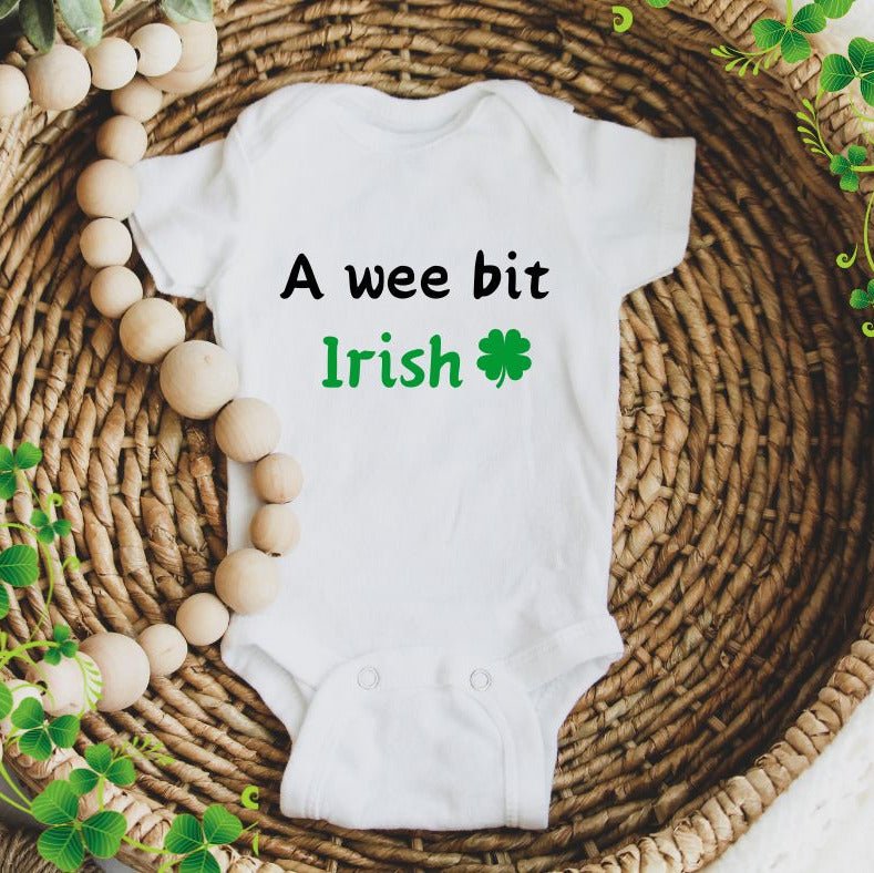 White baby bodysuit with  "we bit Irish" on front and clover sitting in a wicker bowl with beads. Little Gift Nook