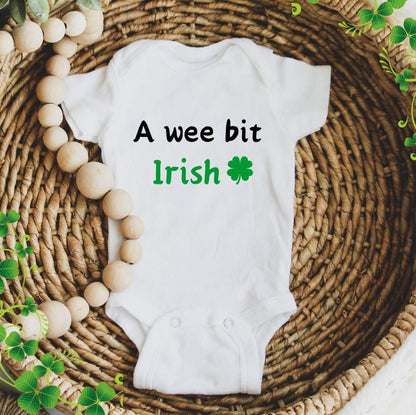 White baby bodysuit with  "we bit Irish" on front and clover sitting in a wicker bowl with beads. Little Gift Nook