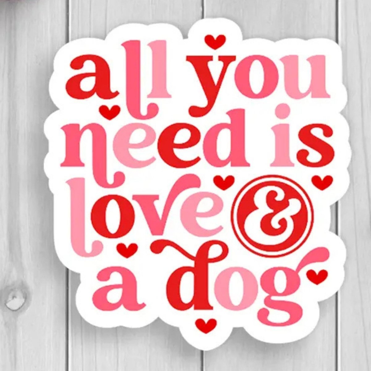 red pink text sticker "all you need is love & a dog" with hearts with white background sitting on a wood background. 