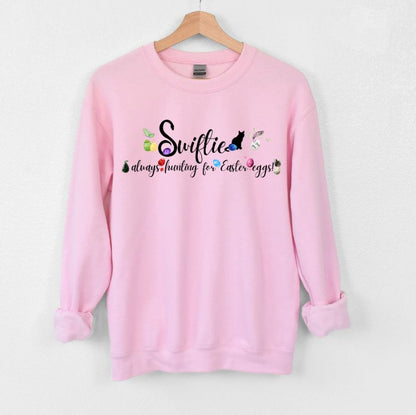 Pink crewneck sweatshirt with Swiftie always hunting for Easter Eggs in black lettering and colored Easter eggs with patterns and items associated with each era.  Little Gift Nook.