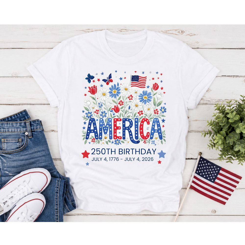 White t-shirt with 'America 250th Birthday' floral design on front laying on wooden surface with jeans and an American flag.