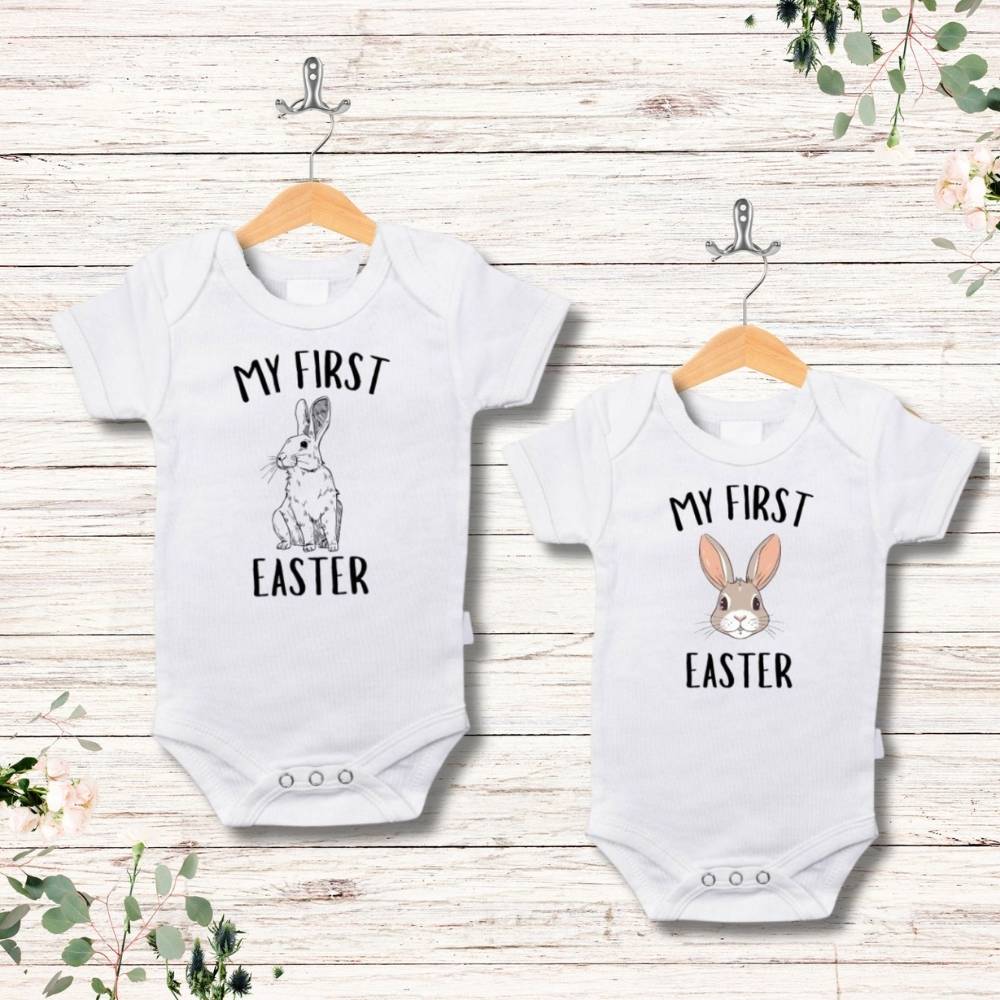 Two white onesies with "My first Easter" on front and a bunny image hanging on a faux wood background.