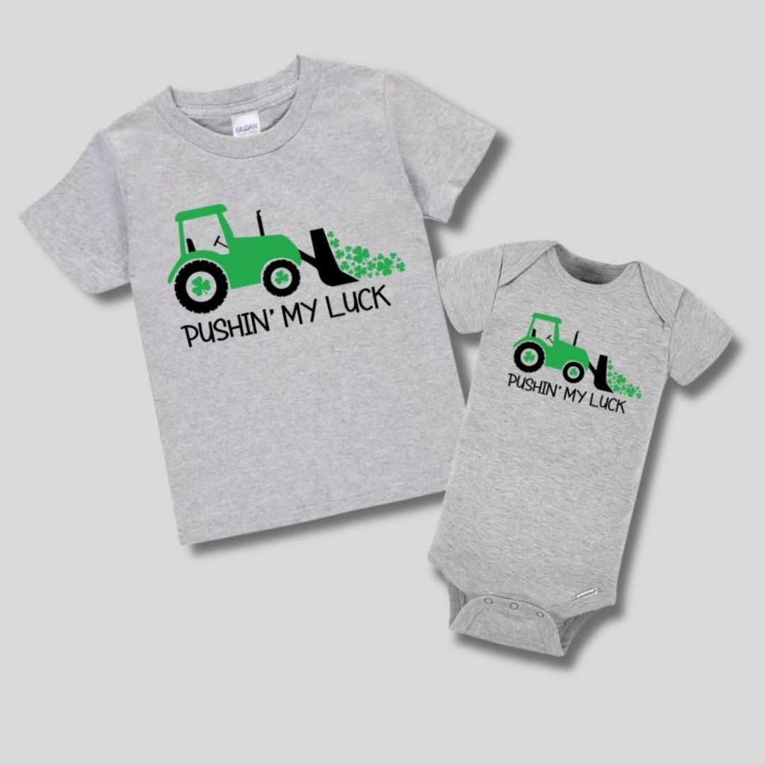 Gray baby onesie and kids tshirt with green tractor and clovers "pushing my luck" tshirts. On gray background. Little Gift Nook