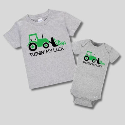 Gray baby onesie and kids tshirt with green tractor and clovers "pushing my luck" tshirts. On gray background. Little Gift Nook