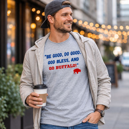 A man wearing a light gray heather t-shirt with a blue and red Buffalo, quote "Be Good, Do Good, God Bless, and Go Buffalo" on front, styled with an open beige zip-up jacket and a dark baseball cap, holding a coffee cup in an outdoor urban setting.