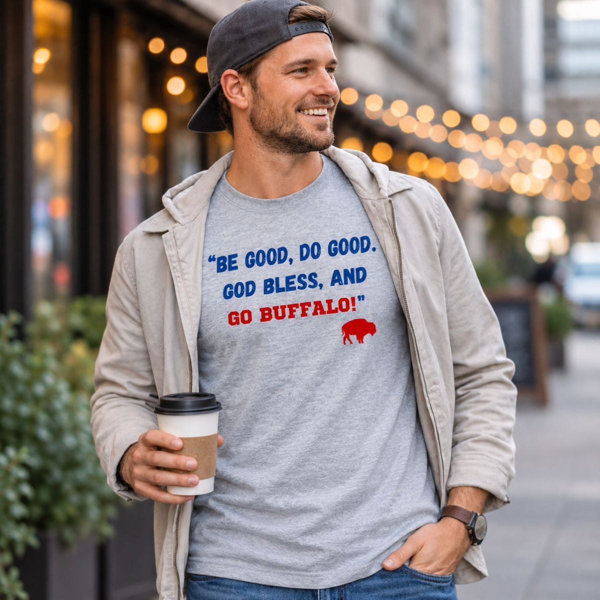 A man wearing a light gray heather t-shirt with a blue and red Buffalo, quote "Be Good, Do Good, God Bless, and Go Buffalo" on front, styled with an open beige zip-up jacket and a dark baseball cap, holding a coffee cup in an outdoor urban setting.