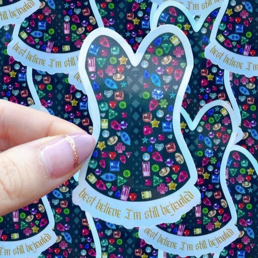 Colorful glittery bodysuit sticker with text "best believe I'm still bejeweled" held by a hand against a multicolored background