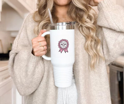 Woman holding white tumbler with pink ribbon image sticker with "best MOM ever" on front sitting on gray background. Little Gift Nook