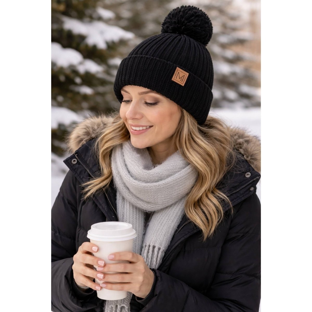 Woman wearing a black knit beanie with a pom-pom and holding a coffee cup outdoors in winter.