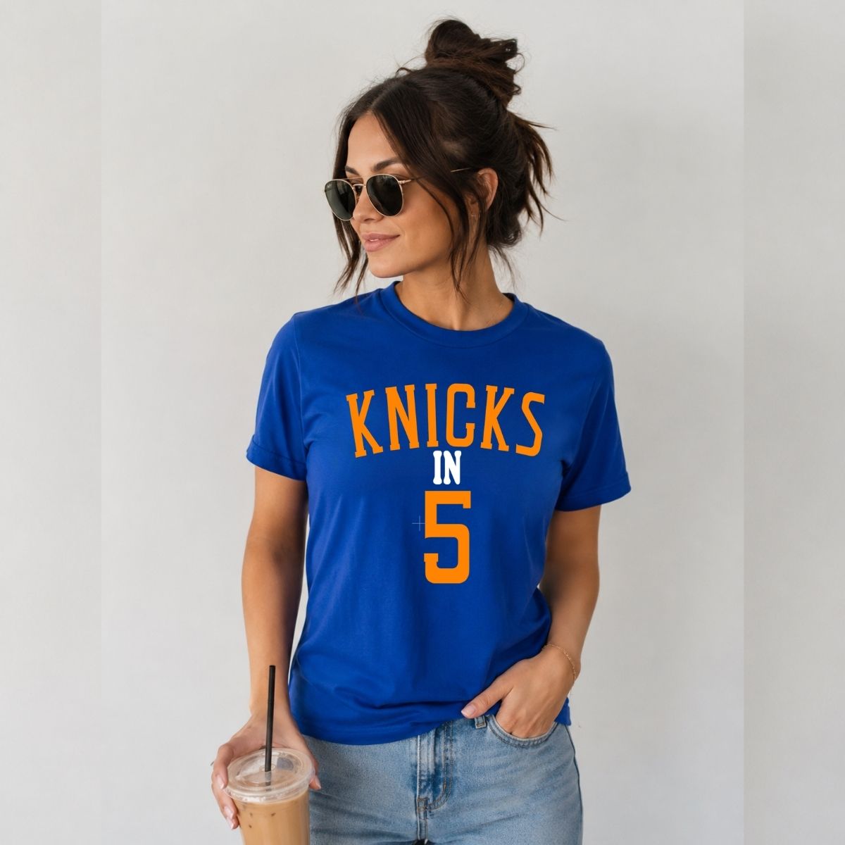 Woman wearing royal blue and orange "Knicks in 5" tshirt holding latte on a gray background.