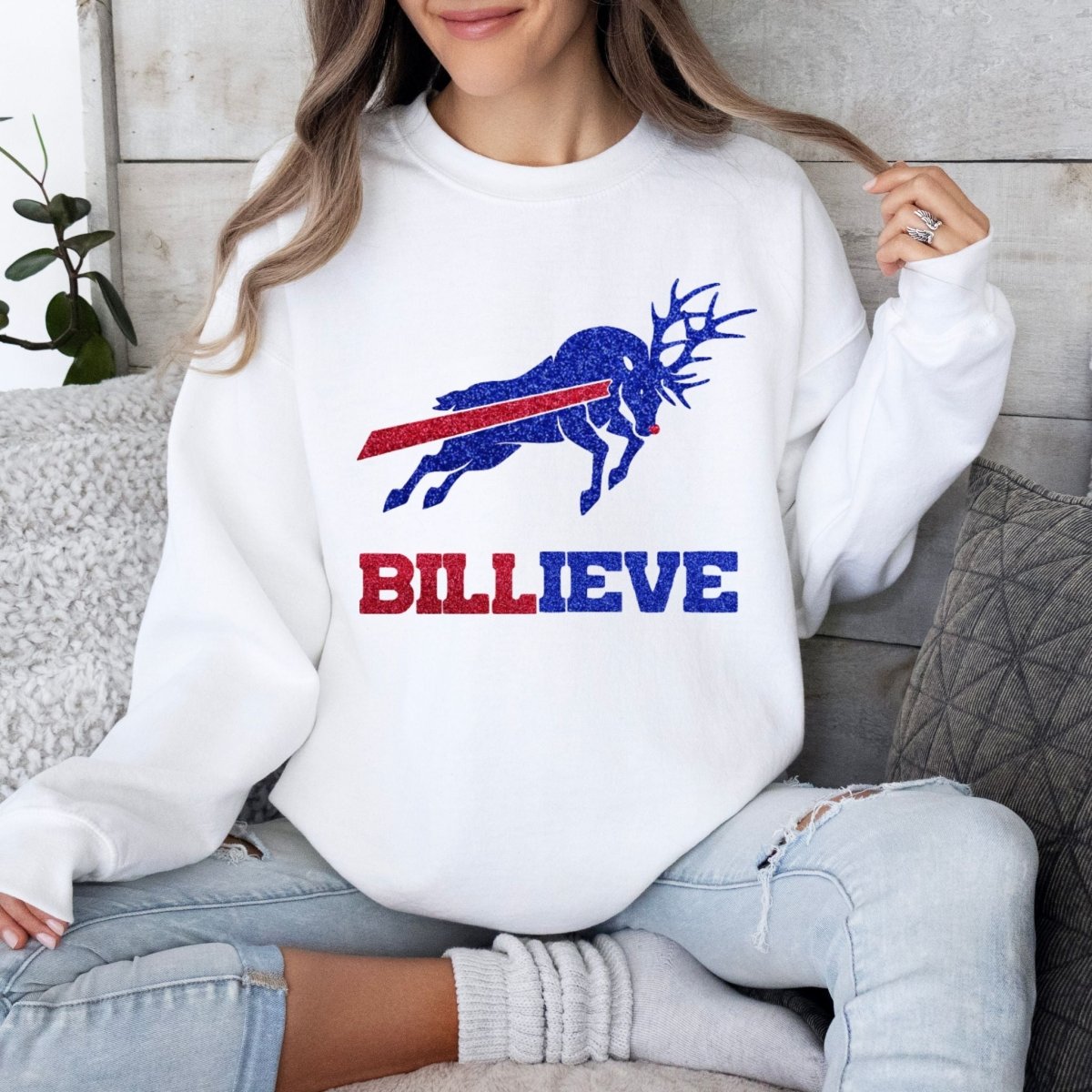 woman wearing white sweatshirt with blue and red glitter buffalo football theme reindeer and BILLEVE under it sitting on gray couch with pillows.
