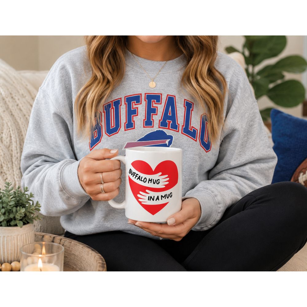 Woman in Buffalo sweatshirt holding Buffalo Hug in a Mug white ceramic coffee mug with red heart design, seated on couch in cozy home setting.