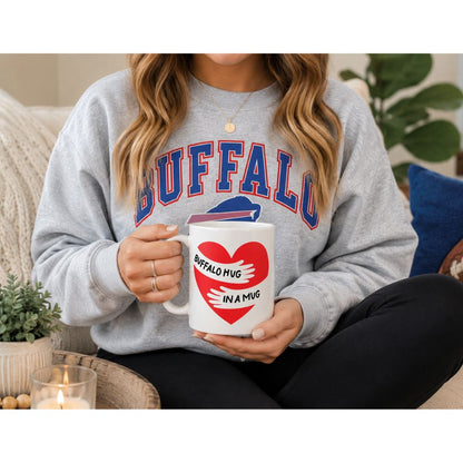Woman in Buffalo sweatshirt holding Buffalo Hug in a Mug white ceramic coffee mug with red heart design, seated on couch in cozy home setting.